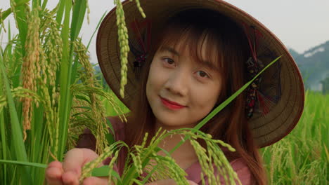 Vietnamese-farmer-young-female-woman-wearing-traditional-bamboo-hat-showing-at-camera-rice-crop-in-field-plantation-,-close-up-portrait