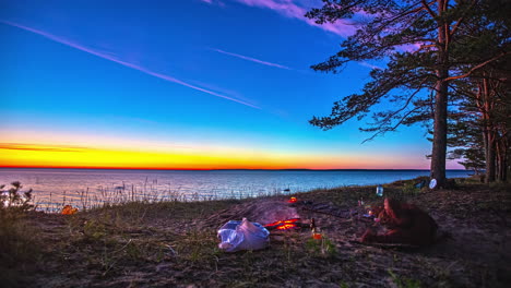 Group-of-people-camping-near-seaside-with-burning-bonfire,-magical-sunset-sky