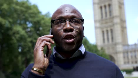 Professional-bald-man-with-glasses-talking-on-the-phone-outside-in-a-park-setting-on-a-sunny-day