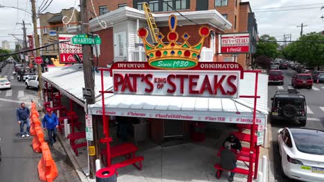 Pat’s-King-of-Steaks-in-Philadelphia,-busy-intersection-at-Wharton-St,-people-walking,-cars-passing-by,-aerial