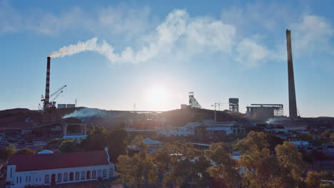 Aerial-Track-back,-Mount-Isa-mine-chimney-stacks-with-plumes-of-smoke-and-steam-coming-out-of-them-on-a-blue-sky-background