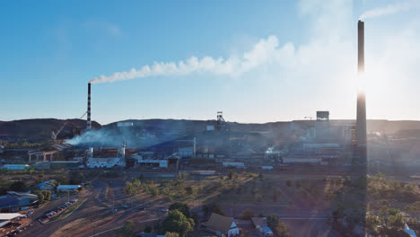 Aerial-Slow-motion,-Track-Back,-Chimneys-from-Mount-Isa-mines-with-puffing-smoke-against-a-sunset-background-and-blue-sky