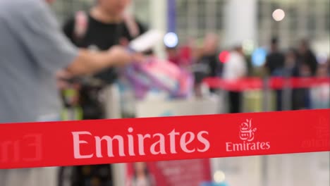 El-Logotipo-De-Emirates-Airlines,-La-Aerolínea-De-Los-Emiratos-Árabes-Unidos,-Se-Ve-En-Una-Cinta-De-Barrera-Mientras-Los-Pasajeros-Del-Vuelo-Hacen-Fila-Para-Registrarse-En-El-Aeropuerto-Internacional-Chek-Lap-Kok.