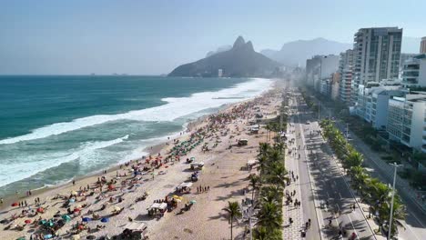 Stunning-drone-footage-over-Rio-de-Janeiro's-Ipanema-Beach-showcasing-the-busy-beach,-ocean-waves,-majestic-mountains,-city-skyline,-and-traffic-on-the-road