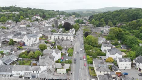 Aerial-tracking-shot-of-black-car-driving-through-scenic-UK-town-surrounded-by-greenery-and-houses,-Devon,-UK