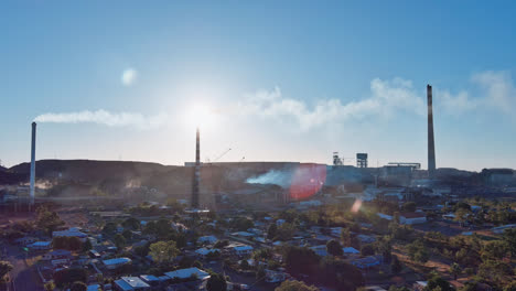 Aerial,-Track-right,-Mount-Isa-town-with-the-chimney-stacks-from-the-mines-in-the-background-with-smoke-in-the-blue-sky-and-lens-flares-from-the-sun