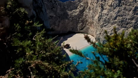 Slow-motion-view-of-Smugglers-Cove-Shipwreck-boat-on-sandy-beach-headland-coastline-with-trees-in-foreground-Navagio-Island-Zakynthos-Greece-Mediterranean-Sea-Europe