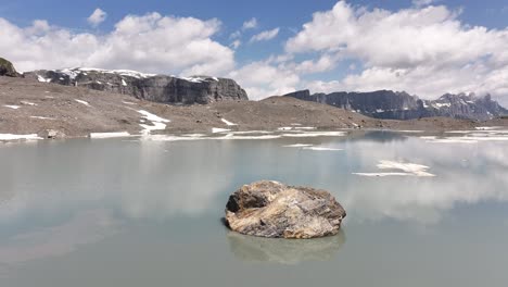 Vista-Aérea-De-Una-Roca-De-Piedra-Solitaria-En-Medio-Del-Lago-Glacial-Gletschersee-En-Klausenpass,-Urner-boden,-Suiza,-Destacando-Tanto-Klausenpass-Como-Griesslisee