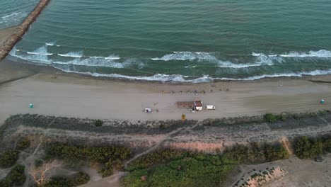 Aerial-view-of-a-calm-wave-beach-at-sunset-with-a-beach-bar,-umbrellas,-sun-loungers,-portable-toilets,-and-a-van-on-the-shore