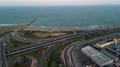 Aerial-view-of-a-coastal-highway-with-traffic,-adjacent-to-the-sea-and-industrial-buildings-in-a-complex,-showcasing-infrastructure-near-nature-with-vegetation-and-trees