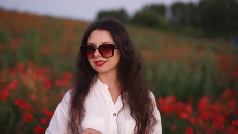 beautiful-dark-haired-girl-in-a-field-of-wildflowers-and-red-poppies,-wearing-a-dress-and-smiling-while-putting-on-sunglasses