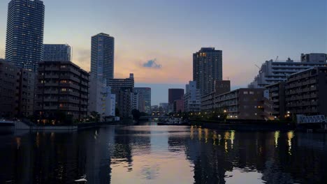 Eine-Ruhige-Stadtlandschaft-In-Der-Abenddämmerung-Mit-Spiegelungen-Der-Wolkenkratzer-Auf-Einem-Ruhigen-Fluss