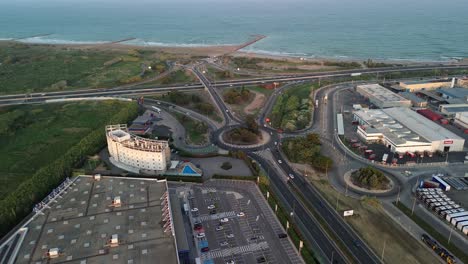 Aerial-view-of-a-coastline-with-a-large-circular-building,-roads,-vehicles,-and-a-parking-lot,-showcasing-the-interaction-between-infrastructure-and-nature