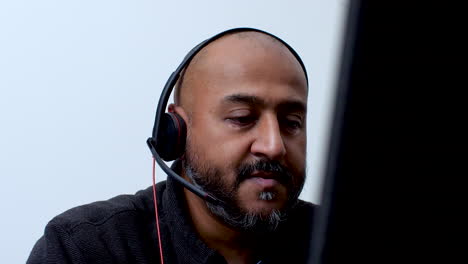 Close-up-of-a-bald-South-Asian-man-wearing-a-headset,-talking-while-working-remotely-from-home