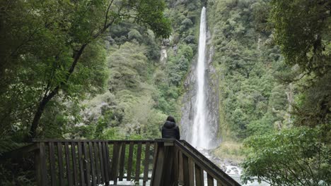 Joven-Caucásica-Con-Una-Chaqueta-Caminando-Y-Mirando-Una-Cascada-En-Un-Bosque-En-Thunder-Creek-Falls,-Costa-Oeste,-Nueva-Zelanda