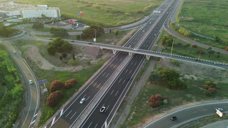 Luftaufnahme-Einer-Autobahn-Mit-Einer-Brücke-Mit-Vegetation-Und-Einem-Gewerbegebiet-Auf-Der-Anderen-Seite-Mit-Autoverkehr-Bei-Sonnenuntergang