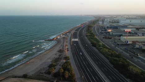 Aerial-view-of-a-coastal-road-with-multiple-lanes-next-to-a-beach,-with-buildings-on-the-other-side,-showing-the-interaction-between-infrastructure-and-nature