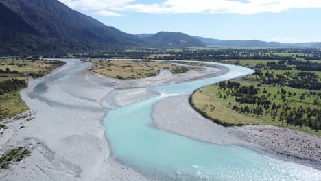 Drone-view-of-blue-river-with-glacier-water-flowing-in-between-fields-at-West-Coast,-New-Zealand