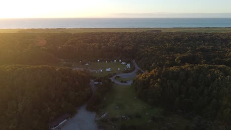 Drone-view-of-a-the-ocean-and-a-campground-with-campervans-in-between-forests-during-sunset-at-Lake-Mahinapua,-Hokitika,-West-Coast,-New-Zealand