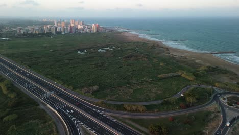 Vista-Aérea-De-Un-Paisaje-Urbano-Costero-Al-Atardecer,-Con-Una-Autopista-Y-Una-Rotonda-En-Primer-Plano-Y-Edificios-Y-Naturaleza-Al-Fondo-Cerca-De-La-Playa