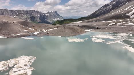 Imagen-Aérea-De-Un-Dron-Volando-Lentamente-Sobre-Una-Laguna-Cristalina-Que-Refleja-Las-Montañas-Y-El-Cielo-En-Medio-De-Los-Alpes-Suizos