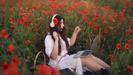 Schönes,-Fröhliches-Dunkelhaariges-Mädchen-Mit-Einer-Mohnblume-Im-Haar,-Sitzt-In-Einem-Feld-Aus-Wildblumen-Und-Roten-Mohnblumen,-Trägt-Ein-Kleid,-Lächelt,-Tanzt-Und-Singt-Mit-Kabellosen-Kopfhörern