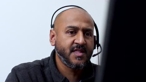 Bald-man-with-a-short-beard-is-wearing-a-headset-and-talking-while-looking-at-a-computer-screen,-possibly-engaged-in-a-virtual-meeting-or-providing-customer-support