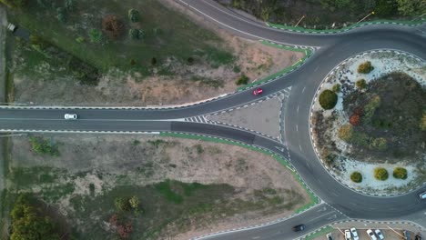 Aerial-tracking-shot-of-a-red-and-white-car-at-a-roundabout-and-on-a-bridge-over-a-highway