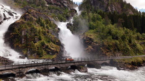 Famous-Låtefossen-waterfall-in-Norway,-aerial-view-car-driving-across-old-stone-bridge