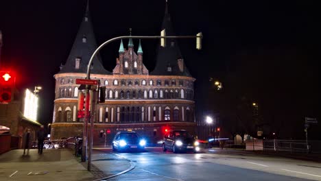 Zoom-out-time-lapse-of-a-busy-night-intersection-in-front-of-the-Museum-Holstentor-western-gate-in-Lübeck,-Germany