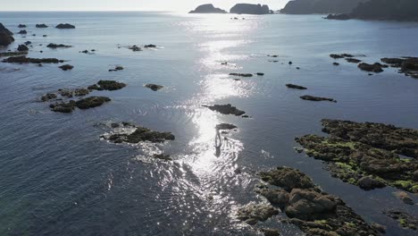 Aerial-view-of-Stand-Up-Paddle-boarding-on-calm-water-with-sun-reflecting-along-the-rocky-coastline-in-the-South-of-Ireland-on-a-calm-day
