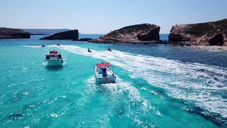 Drone-following-boats-and-men-jet-skiing-on-crystal-clear,-turquoise-water-on-comino-island-in-malta