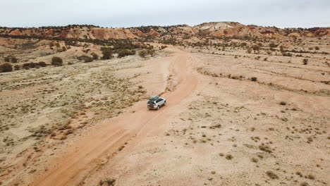 Aerial-tracking-offroad-vehicle-on-remote-dirt-road-leading-into-colorful-desert-hills