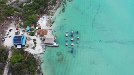 Aerial-Birds-Eye-View-of-Seaside-Shack,-House-with-Solar-Panels-on-Shore-and-Umbrella-Chairs-in-Beautiful-Blue-Water