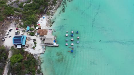 Luftaufnahme-Aus-Der-Vogelperspektive-Einer-Hütte-Am-Meer,-Eines-Hauses-Mit-Sonnenkollektoren-Am-Ufer-Und-Sonnenschirmen-Im-Wunderschönen-Blauen-Wasser