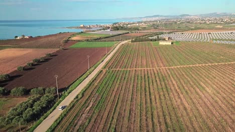 Aerial-footage-of-a-white-car-driving-on-the-village-road,-through-the-fields-and-greenhouses