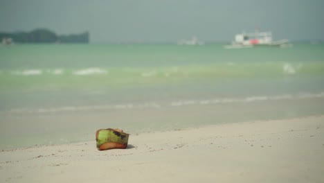 Cáscara-De-Coco-En-La-Orilla-Del-Mar-Con-Hermosas-Olas-Chapoteando-Suavemente-En-La-Famosa-Isla-De-El-Nido,-Filipinas,-Durante-El-Día-Soleado---Toma-Amplia