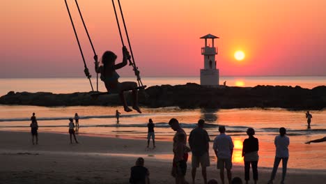 Mädchen-Schaukelt-Auf-Einer-Schaukel,-Leute-Schauen-Auf-Den-Leuchtturm-Und-Den-Sonnenuntergang-Am-Strand,-Khao-Lak,-Thailand