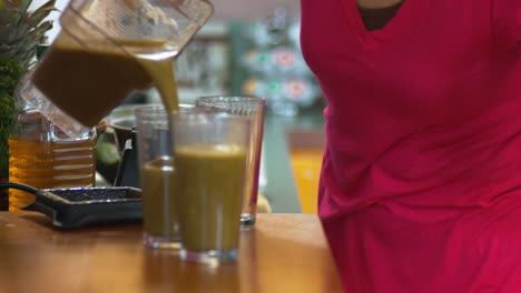 A-female-hand-holding-a-recently-blended-green-smoothie-pours-them-in-to-three-glasses-on-kitchen-counter