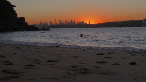 Sunset-over-Sydney-skyline-from-Lady-Bay,-red-sky-bushfire-smoke,-silhouette-of-woman-swimming-and-rock-fishermen---rack-focus,-4K