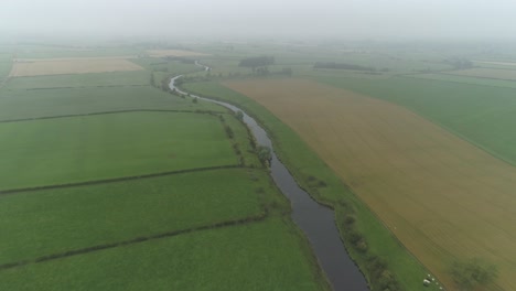 Toma-Aérea-Sobre-Las-Tierras-De-Cultivo-Y-El-Río,-Siguiendo-El-Río-En-Un-Día-Brumoso