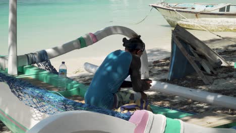 Asiatischer-Mann-Sitzt-Auf-Einem-Boot-An-Einem-Sandstrand,-Macht-Eine-Pause-Und-Schaut-Auf-Sein-Smartphone-Auf-Gili-Air,-Indonesien