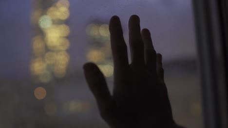 Hand-entres-frame-and-rests-on-glass-window-overlooking-city-night