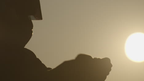 Hand-held-slow-motion-shot-of-male-silhouette-wearing-a-car-racing-helmet-putting-on-thick-gloves-with-sun-setting-in-the-background