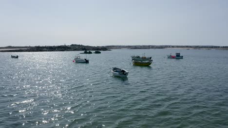 Low-drone-footage-over-small-fishing-boats-in-peaceful-bay-at-anchor-approaching-rocks-with-shoreline-and-church-in-the-background,-shot-in-Guernsey