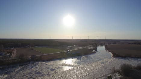 Drohnenaufnahmen-Eines-Zugefrorenen-Finnischen-Flusssees-Mit-Wunderschönem-Sonnenuntergang-Und-Windmühlen-Am-Horizont