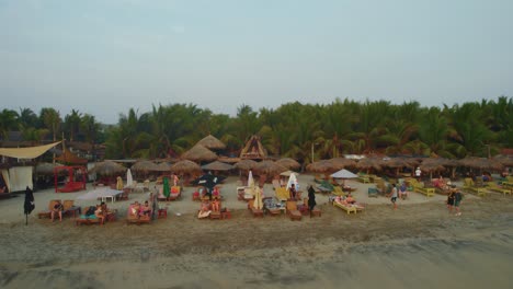 Aerial-view-of-people-relaxing-next-to-palm-trees-by-the-ocean,-in-a-beach-club-in-Puerto-Escondido,-Mexico