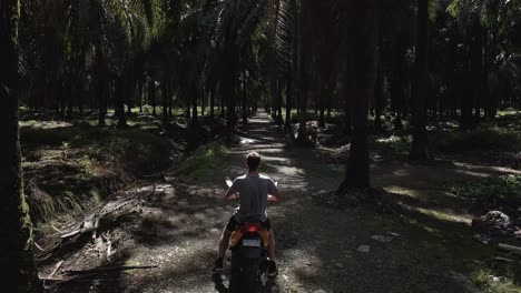 Young-man-walking-towards-a-scooter,-then-driving-off-through-a-dark-palm-tree-forest-in-Costa-Rica