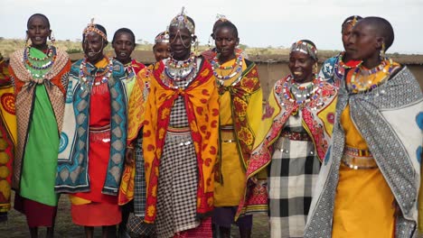 Eine-Zeitlupenaufnahme-Des-Tanzes-Der-Frauen-Der-Massai-Stämme-Im-Masai-Mara-Nationalpark-In-Kenia-Am-Späten-Nachmittag,-Während-Sie-Ihre-Traditionelle-Kleidung-Und-Ihren-Schmuck-Mit-Lehmhäusern-Präsentieren