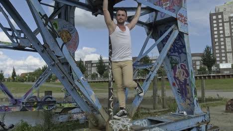 Flexible-male-dancer-in-casual-wear-swinging-legs-in-air-while-practicing-on-metal-construction-and-looking-at-camera